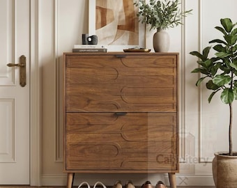 May include: A medium-brown wooden shoe cabinet featuring two drop-down doors with a carved geometric arch pattern and slim black handles. The cabinet stands on four tapered legs, with several pairs of shoes placed neatly underneath.