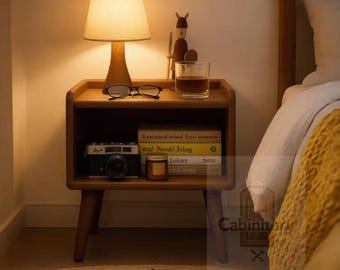May include: A wooden nightstand with a lamp, glasses, a glass of liquid, a camera, and a stack of books. The lamp has a beige shade and emits a warm glow. A brown slipper rests on the floor in front of the nightstand, which features four legs.