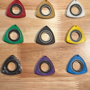 Puede incluir: Nueve spinners triangulares de colores, rojo, amarillo, blanco, verde, negro, amarillo, negro, morado y azul. Cada spinner tiene un rodamiento central y un agujero en la parte superior para un cordón. Los spinners están dispuestos sobre una superficie de madera.