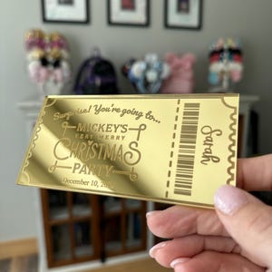 May include: A rectangular gold-toned mirror-finish ticket featuring the engraved text: Surprise! You're going to... MICKEY'S VERY MERRY CHRISTMAS PARTY, December 10, 2026, and the name Sarah, alongside a vertical barcode graphic on the right side.