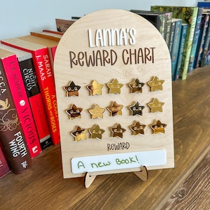 May include: A wooden reward chart with the name "Lanna's" and the text "Reward Chart" in brown lettering. The chart has 12 gold star stickers with smiley faces on them. The reward is written on a white dry erase board and says "A new book!"