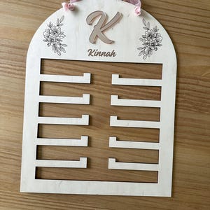 May include: A wooden, arched, personalized jewelry holder with the letter "K" and the name "Kinnah" engraved on it. The holder has eight slots for jewelry and is decorated with floral designs.