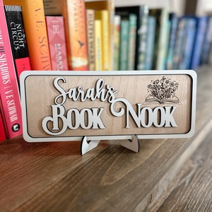 May include: A wooden sign with the text "Sarah's Book Nook" in white lettering. The sign has a decorative design of a book with flowers growing out of it. The sign is on a stand and in front of a bookshelf.