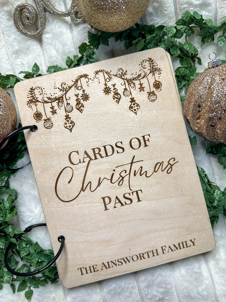以下が含まれることがあります： クリスマスのガーランドと「Cards of Christmas Past」と「The Ainsworth Family」の文字で飾られた、リングバインダー付きの木製ボックスです。