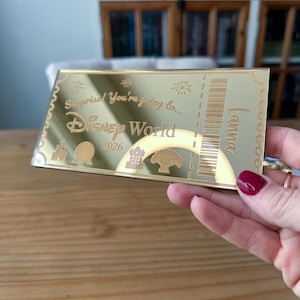 May include: A rectangular gold-toned mirror ticket featuring the engraved text Surprise! You're going to... Disney World 2026, the name Lanna, a barcode, and icons of four park landmarks. The ticket has a scalloped border design along the edges.