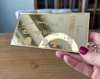 May include: A rectangular gold-toned mirror ticket featuring the engraved text Surprise! You're going to... Disney World 2026, the name Lanna, a barcode, and icons of four park landmarks. The ticket has a scalloped border design along the edges.