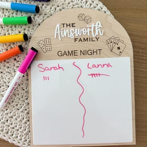 May include: An arched wooden game night scoreboard featuring the text The Ainsworth Family and Game Night, with engraved dice, dominoes, and playing cards. A white dry-erase surface displays the names Sarah and Lanna with pink tally marks.