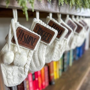 May include: Cream-colored cable knit Christmas stockings with brown leather name tags hang from a wooden beam. Each stocking has a white pom-pom and is personalized with a name. Greenery is placed above the beam, and a row of colorful books is visible below.