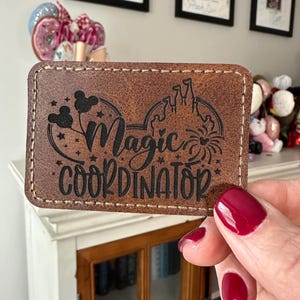 May include: A brown leather patch with the words "Magic Coordinator" in black script. The patch features a Mickey Mouse silhouette and a castle design. The edges are stitched with white thread.