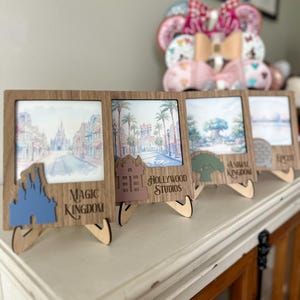 Könnte beinhalten: Vier Holz-Fotorahmen mit Aquarelldrucken von Themenparkszenen. Jeder Rahmen hat ein einzigartiges Ausschnittdesign und den Text: "Magic Kingdom", "Hollywood Studios", "Animal Kingdom" und "Epcot". Die Rahmen stehen auf einer weißen Oberfläche.