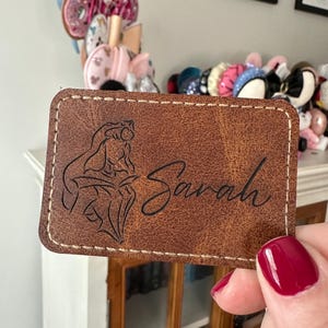 May include: A rectangular brown faux leather patch with rounded corners and light-colored stitching along the edges. The patch features an engraved outline of a female character reading a book next to the name Sarah in a cursive script font.