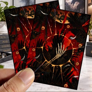 May include: A rectangular trading card featuring a dark illustration of black ravens, floating eyeballs, and a central skeletal hand reaching out from a glowing golden ring against a deep red and black background with scattered white feathers.