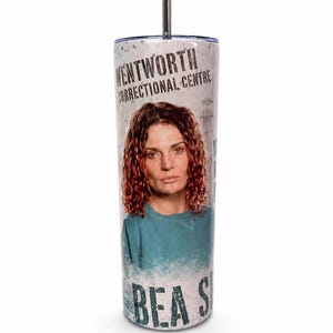 May include: A cylindrical tumbler with a metal straw featuring a graphic print of a person with curly reddish-brown hair wearing a teal shirt. The text reads Wentworth Correctional Centre and Bea S. The design includes a lighthouse illustration.