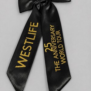 Könnte beinhalten: Eine schwarze Haarschleife aus Satin mit zwei langen Bändern. Auf dem linken Band steht der goldene Glitzertext WESTLIFE, auf dem rechten Band der goldene Glitzertext 25 THE ANNIVERSARY WORLD TOUR. Die Schleife hat eine glatte, leicht glänzende Oberfläche.