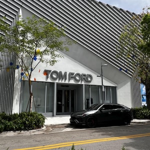 May include: Exterior shot of a Tom Ford store with a modern architectural design. The building has a gray and white facade with the brand name prominently displayed. A black car is parked in front of the store. Trees and greenery frame the scene.