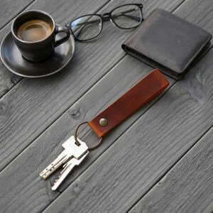 Puede incluir: Un llavero de cuero marrón con un botón de presión metálico y una anilla circular que sujeta dos llaves plateadas, acompañado de una taza de café negra sobre un platillo, gafas de montura negra y una cartera de cuero marrón oscuro.