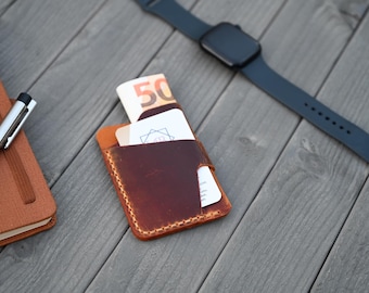 May include: A brown leather card holder with visible stitching containing a fifty euro banknote and a white business card with a geometric logo, positioned near a silver pen, a brown notebook, and a black smartwatch with a silicone band.