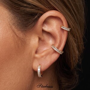 Puede incluir: Tres accesorios dorados para la oreja con piedras transparentes en corte baguette. Un pequeño pendiente de aro se lleva en el lóbulo, mientras que dos pendientes de presión se sitúan en el cartílago superior. El texto Starbussss es visible abajo.