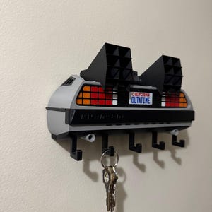 May include: A gray and black key holder shaped like the DeLorean time machine from Back to the Future. It has a license plate that reads "OUTATIME" and includes five key hooks. The key holder is mounted on a white wall.