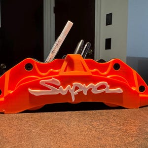 May include: A bright orange desk organizer shaped like a car brake caliper with the word Supra embossed in white on the front. The organizer features multiple slots for holding pens and markers, with a white pen visible inside one of the openings.