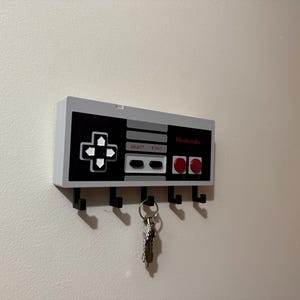 Op de afbeelding: Een aan de muur gemonteerde sleutelhouder ontworpen als een klassieke Nintendo-controller. De sleutelhouder is wit, zwart en grijs, met rode accenten en het Nintendo-logo. Het heeft vijf zwarte haken voor sleutels.