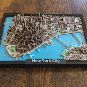 Peut inclure: Une carte en relief 3D encadrée de New York avec des bâtiments et des infrastructures miniatures marron sur un fond d'eau bleu vif. Le cadre noir comporte une étiquette en bas avec le texte New York City écrit en lettres blanches.