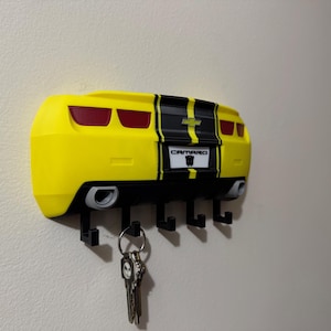 May include: A yellow and black Camaro-themed key holder mounted on a wall. The key holder is shaped like the rear of a car, with red taillights, exhaust pipes, and the word "Camaro" on the license plate. Several black hooks hang below to hold keys.