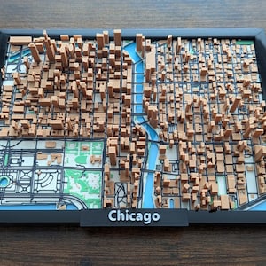 Op de afbeelding: Een ingelijste 3D-kaart van Chicago met beige miniatuurgebouwen en wolkenkrabbers op een platte ondergrond met blauwe waterpartijen en groene parkgebieden. Het zwarte frame bevat een plaatje met het woord Chicago in witte letters.
