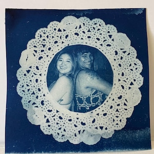 May include: A cyanotype print featuring two people framed by a white lace doily. The subjects are smiling, with one person's arm around the other. The background is a deep blue.