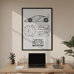 May include: Black framed print featuring a line drawing of a sports car from side, top, and front views. The car has a spoiler and detailed wheels. The print is displayed above a wooden desk with a laptop, plant, and accessories.
