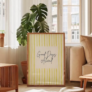 May include: A framed print with a wooden frame featuring the text "Good Days Ahead" in a cursive font. The background has vertical yellow stripes. The print is displayed in a well-lit living room with a large plant.