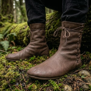 Peut inclure: Bottes en cuir marron avec un laçage et des coutures latérales. Les bottes ont un bout arrondi et une hauteur mi-mollet. Les bottes sont présentées en extérieur sur de la mousse et des feuilles vertes.
