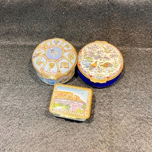 以下が含まれることがあります： ヒンジ付きの蓋が付いた3つの装飾用小物入れ。1つはライトブルーの歴史的な年表デザインの丸い箱。もう1つは青いベースに鳥と花が描かれた丸い箱。最後は大きな山が描かれた風景画の長方形の箱です。
