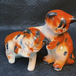 Può includere: Una statuetta in ceramica raffigurante due gattini calico con macchie arancioni, bianche e nere. Un gattino è seduto in posizione eretta mentre l'altro è sdraiato, appoggiando la testa contro il primo. Entrambi hanno tratti del viso dipinti.
