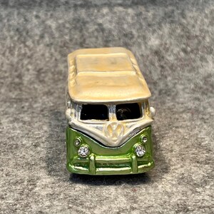 May include: A miniature metal van figurine featuring a cream-colored roof and a metallic green body. The front displays a white V-shaped emblem, two clear crystal headlights, and a windshield with dark interior detailing.