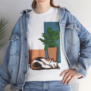 May include: White t-shirt featuring a graphic of a calico cat lying down, a potted plant, and geometric shapes in brown and teal. The cat has patches of black, brown, and white fur. The shirt is worn with a light blue denim jacket.