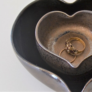 May include: Two heart-shaped ceramic dishes, one nested inside the other. The smaller dish is a light brown colour with a dark brown rim. The larger dish is a dark grey colour with a metallic sheen. A gold ring with a decorative design is sitting in the smaller dish.