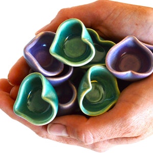 May include: A collection of small, heart-shaped ceramic bowls in shades of green and purple. The bowls are arranged in a pile and held in the hands of a person.