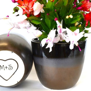 May include: A black and brown ceramic flower pot with a blooming Christmas cactus. The pot is next to a matching vase with a heart-shaped design and the initials "M+S". The cactus has red, white, and pink flowers.