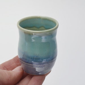 May include: A small, hand-thrown ceramic cup with a light blue glaze that fades to a darker blue at the bottom. The cup has a rounded shape and a slightly flared rim.