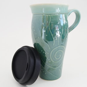 May include: A tall, teal ceramic travel mug with a handle and a black lid. The mug features a decorative swirl and dot design. The lid is off to the side. The mug is on a white background.