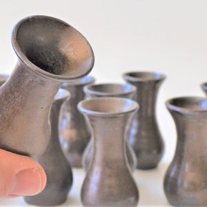 May include: A set of eight small, dark brown ceramic vases. The vases are all the same size and shape, and they have a slightly flared opening.