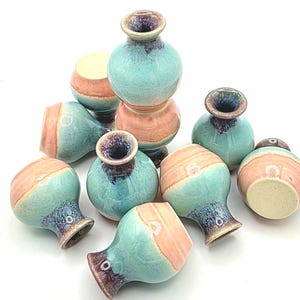 May include: A collection of small, handmade ceramic vases. Each vase features a turquoise glaze on the body, with a band of peach-colored glaze around the middle. The rims and interiors have a dark, speckled finish.