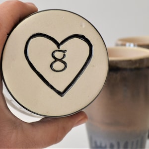 May include: A white ceramic mug with a black heart design and the number 8 inside. The mug is being held by a hand.