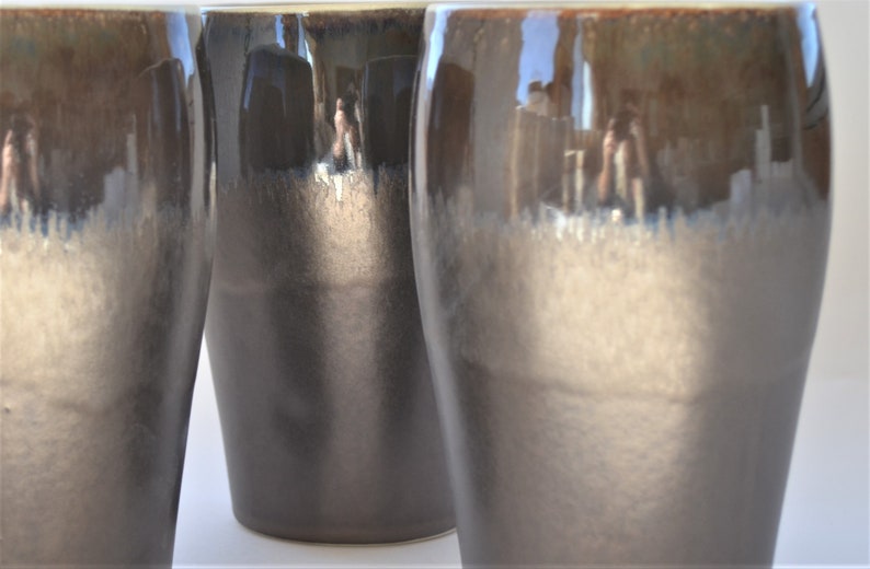 May include: Three ceramic mugs with a dark brown to light brown gradient glaze. The mugs are tall and cylindrical with a rounded bottom.