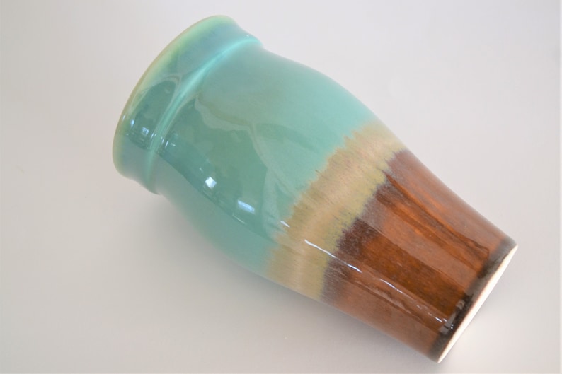 May include: A ceramic vase with a glossy finish. The top section is a light turquoise color, transitioning to a tan band, and then to a rich brown at the base. The vase has a slightly flared rim.