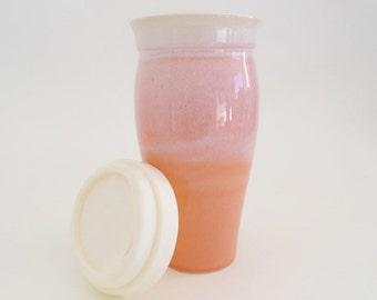 May include: A pink and white ceramic travel mug with a white lid. The mug has a gradient design, fading from white at the top to pink at the bottom.