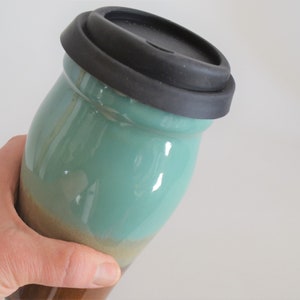 May include: A ceramic travel mug with a black silicone lid. The mug has a teal and brown glaze. The mug is held in a hand. The mug is designed to keep beverages hot or cold.