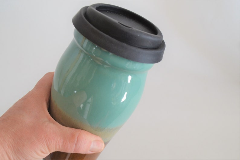 May include: A ceramic travel mug with a black silicone lid. The mug has a teal and brown glaze. The mug is held in a hand. The mug is designed to keep beverages hot or cold.