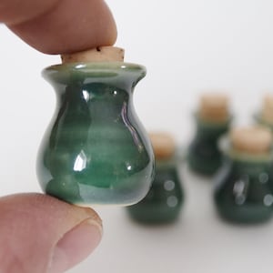 May include: A set of small, green ceramic jars with cork lids. The jars are glazed in a dark green color with a lighter green stripe around the middle. The jars are perfect for storing small items like spices, herbs, or jewelry.